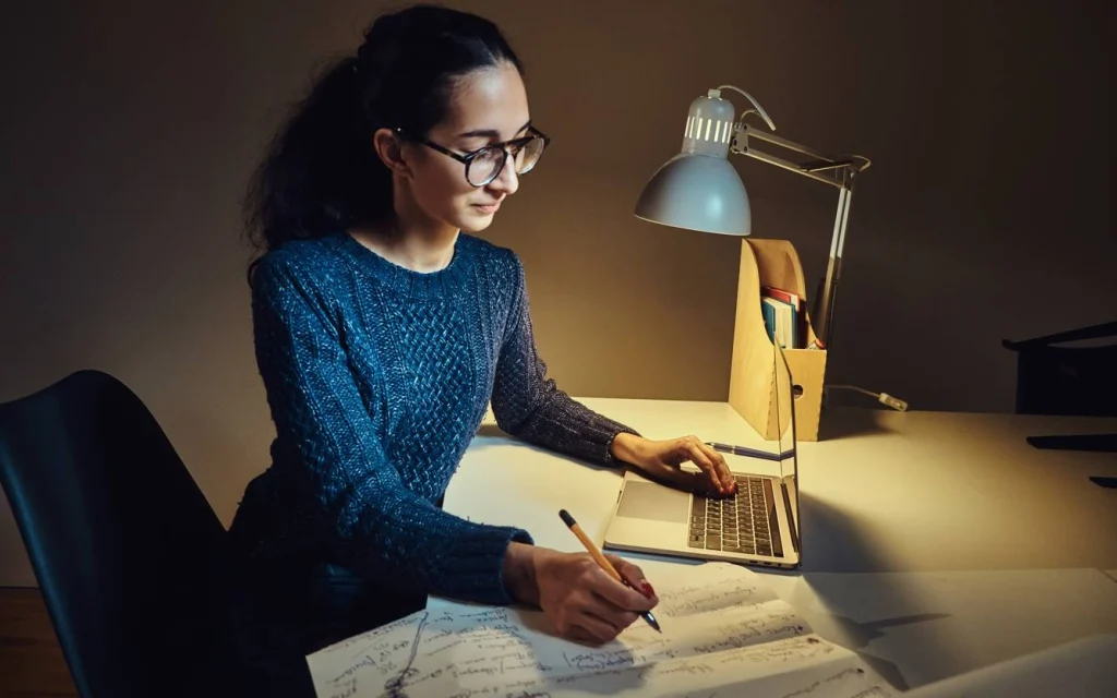 Woman writing notes beside laptop at night